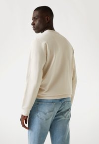 Sudadera beige de manga larga, con cuello redondo y corte relajado, combinada con jeans de color azul claro que tienen bolsillos traseros.
