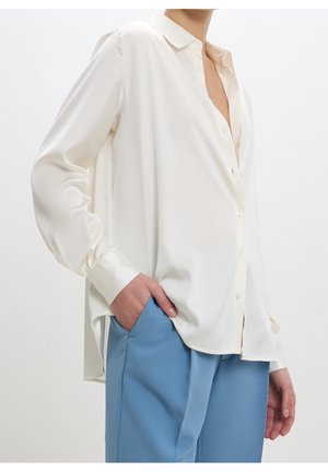 Personne portant une chemise blanche ample à boutons, une main dans la poche d’un pantalon bleu clair taille haute sur un fond uni.