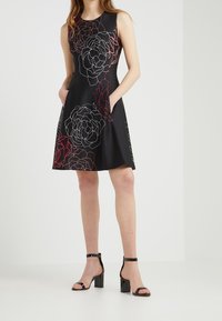 Svart ärmlös klänning med vitt och rött blommönster, med sidofickor och en utställd kjol. Modellen bär svarta klacksandaler med blockklack.