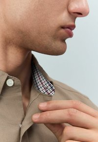 Camisa de manga corta de color caqui con un cuello a cuadros en rojo y azul. Cuenta con cierre de botones y una textura de tela suave.