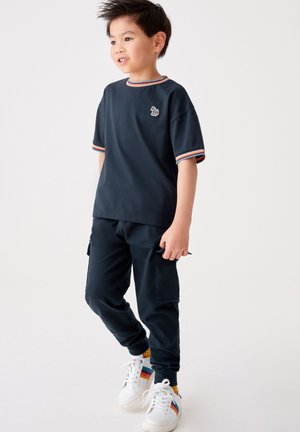 Mørkeblå t-shirt med stribet halsudskæring og logo, parret med matchende joggers. Hvide sneakers med flerfarvede striber.