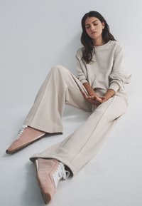 Beež värvi mugavuse komplekt lõdva lõikega; sisaldab pikki varrukaid, laiade sääreosadega pükse ja pehmet kangast. Välimust täiustavad roosad tossud valgete paeltega.