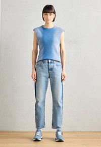 Débardeur côtelé bleu clair, jean bleu classique avec des panneaux latéraux foncés, et baskets bleu avec des accents blancs, posant devant un fond neutre.