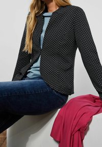 Osoba v černobíle vzorovaném saku, světle modré košili, tmavě modrých džínách, posedávající s růžovou látkou přehozenou přes bílý povrch.