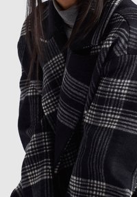 Femme portant un manteau en laine à carreaux noir et blanc par-dessus un pull à col roulé gris, montrant les détails de l'épaule et du haut du torse.