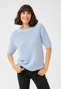 Pull léger bleu à manches courtes, en fil texturé avec un motif de tricot ouvert. Encolure ronde et coupe décontractée, associé à un pantalon sombre.