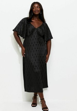 Kobieta w czarnej sukience midi z falbanami na rękawach i delikatnym kwiatowym wzorem, w czarnych szpilkach na kostkę, stoi na tle białej ściany.