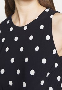 Halenka bez rukávů z námořnického materiálu s bílými puntíky, kulatým výstřihem a mírným nařasením na přední straně. Hladká textura.