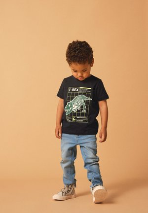 Black cotton t-shirt featuring a green T-Rex skeleton graphic; paired with light blue jeans and grey sneakers with textured soles.
