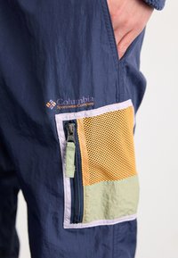 Pantalons imperméables bleus navy avec une poche en mesh aux accents orange et vert, et une fermeture éclair. Logo affiché de manière proéminente.