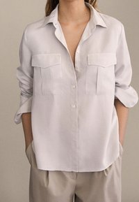 Biała koszula z guzikami z kołnierzykiem, dwoma kieszeniami na piersi i podwiniętymi rękawami; wykonana z gładkiego materiału w luźnym kroju.