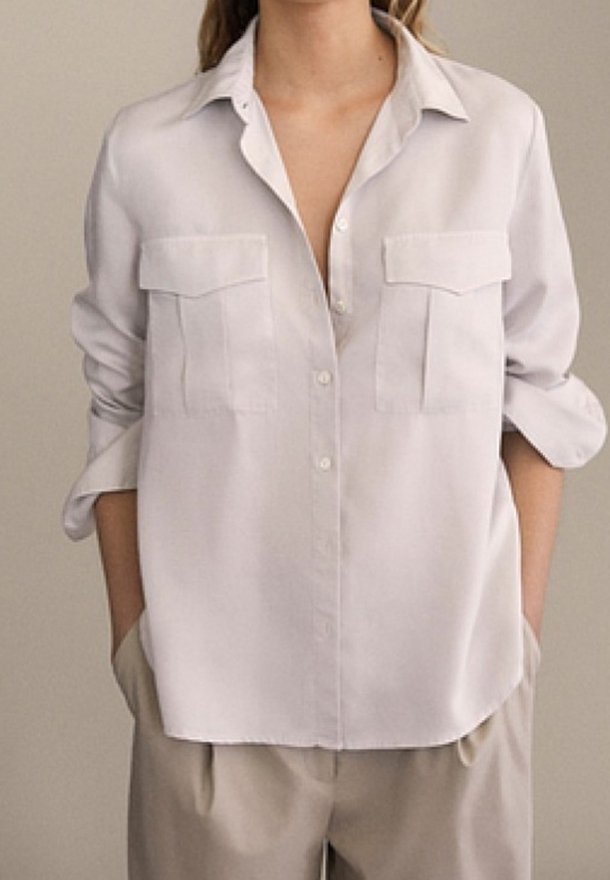 Biała koszula z guzikami z kołnierzykiem, dwoma kieszeniami na piersi i podwiniętymi rękawami; wykonana z gładkiego materiału w luźnym kroju.