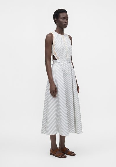 Femme debout portant une robe midi sans manches rayée blanche et bleu clair avec des découpes latérales et des sandales marron sur fond blanc.