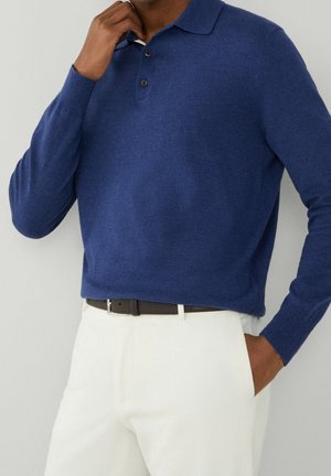 Man draagt een blauw polo-trui met lange mouwen, een witte broek en een zwarte riem, met één hand in zijn zak en de andere bij zijn nek.