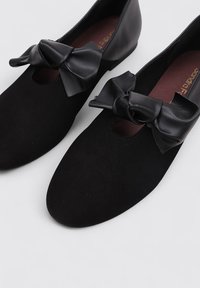Bailarinas de ante negro con punta redonda, que presentan un prominente lazo de satén negro y un detalle troquelado en la parte superior. Acentos de cuero liso.