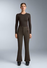 Žena stojí čelom dopredu, oblečená v priliehavom tmavo hnedom tričku s dlhými rukávmi, zodpovedajúcich rozšírených nohaviciach s vreckami na zips a čiernych platformových topánkach.