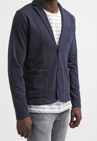 Hombre con blazer azul marino abotonado sobre una camisa blanca con rayas grises y vaqueros gris claro, posando frente a un fondo liso.