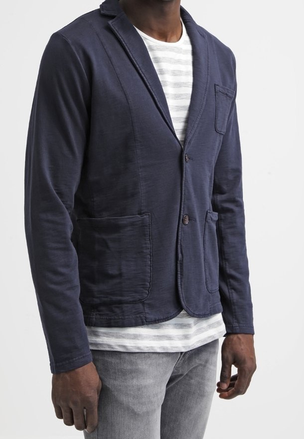 Hombre con blazer azul marino abotonado sobre una camisa blanca con rayas grises y vaqueros gris claro, posando frente a un fondo liso.