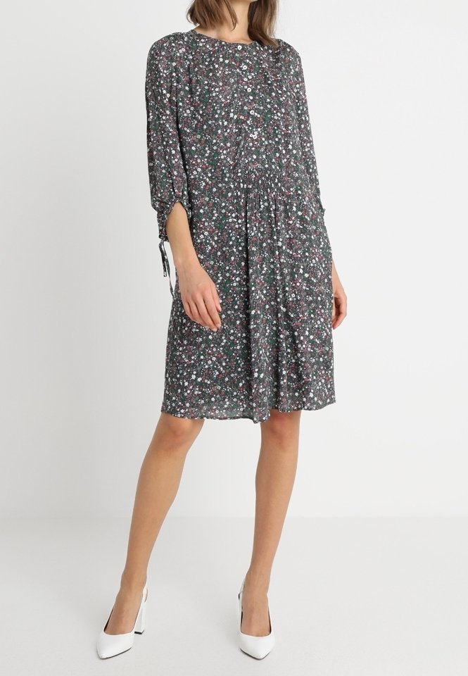 Robe fleurie sombre à manches longues et taille froncée, ornée de petites fleurs roses et blanches sur un fond noir. Modèle portant des talons blancs.