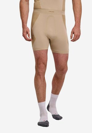 Man draagt beige nauwsluitende shorts en witte sokken met donkergrijze tenen, staat met benen iets uit elkaar op een witte achtergrond.