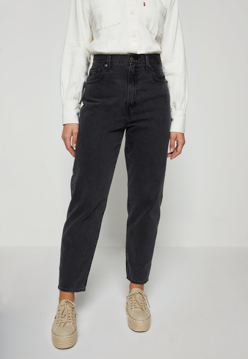 Personne portant un jean noir taille haute associé à des baskets compensées beiges et une chemise blanche boutonnée à manches longues.