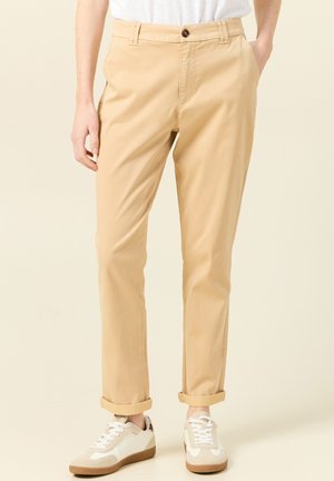 Beige Baumwollhosen mit geradem Bein, Knopfverschluss und umgeschlagenen Säumen, kombiniert mit weißen Sneakers mit braunen Akzenten.