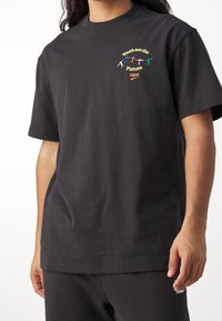 Černé tričko Puma s barevnými postavami, které se drží za ruce, a nápisem "Mládež je budoucnost" na levé horní části hrudi, které nosí osoba.