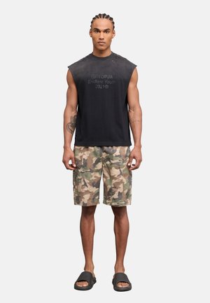 Homme aux cheveux tressés portant un débardeur noir, un short cargo camouflage et des sandales noires, debout devant un fond uni.