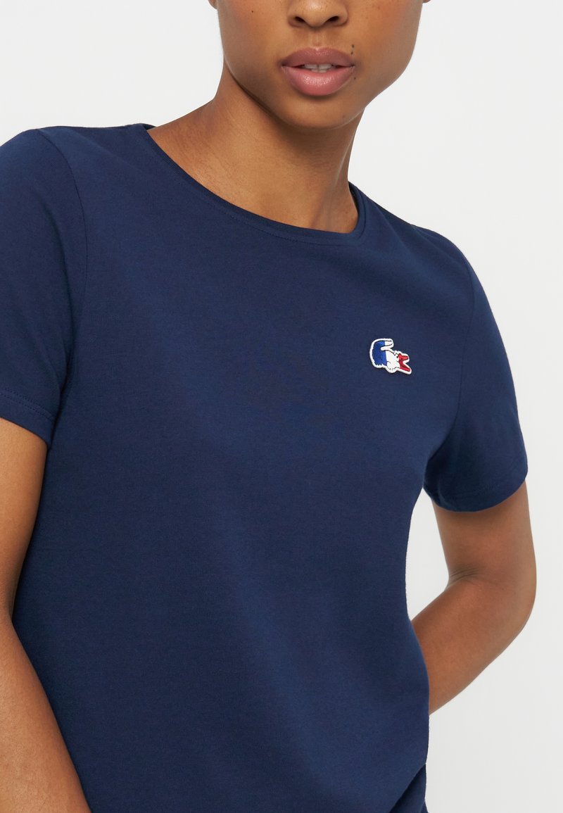 Femme portant un t-shirt bleu marine avec un petit logo brodé d’un crocodile bleu, blanc et rouge sur la poitrine.