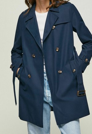 Femme portant un trench-coat bleu marine à double boutonnage avec ceinture et poignets boutonnés par-dessus un top blanc et un jean bleu clair, mains dans les poches.