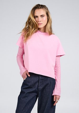 Frau mit langen blonden Haaren, die ein hellrosa kurzärmeliges Oberteil trägt, das über einem strukturierten langärmeligen Hemd und dunkelblauen Jeans liegt.