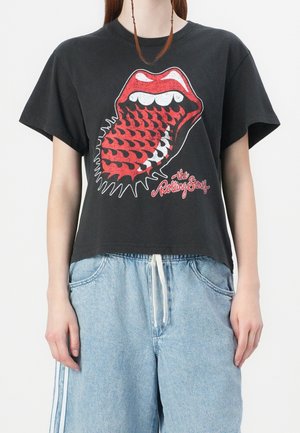 Person iført sort cropped Rolling Stones T-shirt med rød tunge- og læbelogo, kombineret med lyseblå denimshorts med snøre.
