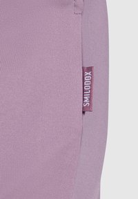 Tejido de color púrpura claro con una textura suave. Presenta una pequeña etiqueta cosida que dice "SMILODOX". Costura mínima visible en la unión.