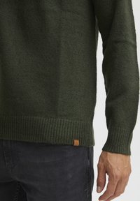 Mørkegrøn strikket sweater med ribbede manchetter og kant, samt et lille læderlogo-patch syet på den nedre kant. Blød tekstur synlig.