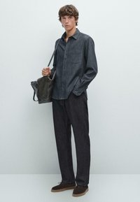 Dunkelblaues Jeanshemd mit Knöpfen, langen Ärmeln und einer Brusttasche, kombiniert mit dunklen Hosen und braunen Wildlederschuhen. Hält eine graue Tasche.