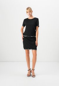 Femme debout, vêtue d'une robe noire avec un passepoil argenté aux manches, à la taille et à l'ourlet, associée à des sandales à talons hauts noir et blanc.