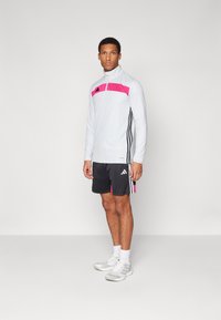 Haut de sport gris clair avec accent rose, shorts noirs avec rayure rose, trois rayures noires sur les côtés et chaussures de sport blanches.