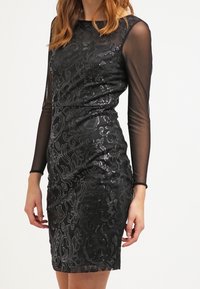 Černé šaty s dlouhými průsvitnými rukávy, zdobené složitými květinovými vzory z flitrů, přiléhavý střih, délka ke kolenům a hladká textura.