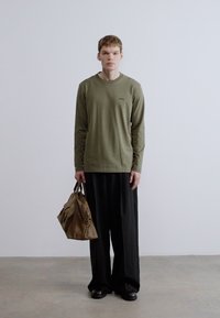 Πουκάμισο μακρυμάνικο σε ελιά πράσινο με μικρό λογότυπο, συνδυασμένο με μαύρο παντελόνι φαρδύ. Το μοντέλο κρατάει μια καφέ τσάντα και φοράει μαύρα παπούτσια.