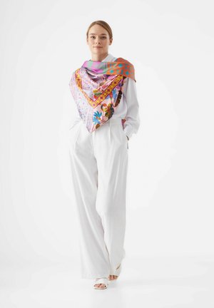 Écharpe en soie, de grande forme carrée, avec des motifs roses et oranges vibrants, des designs floraux et géométriques, portée sur un chemisier blanc et un pantalon.