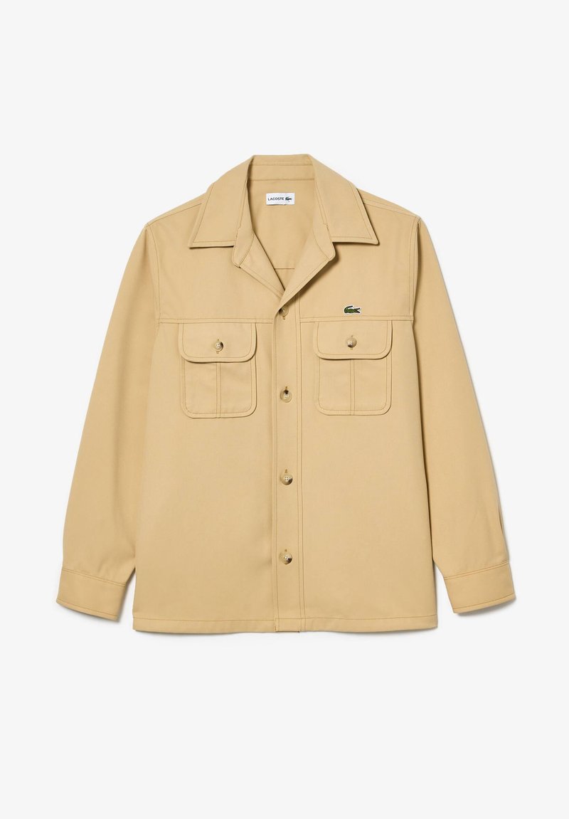 Beige Langarm-Hemdjacke mit Knöpfen, zwei aufgesetzten Brusttaschen und kleinem grünen Krokodil-Logo auf der linken Tasche.