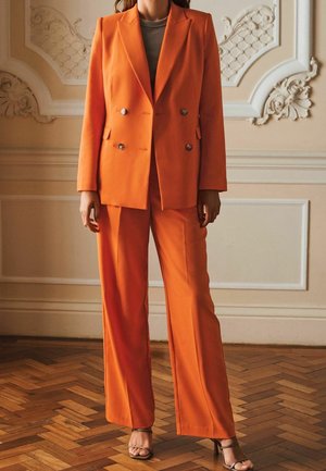 Blazer doppiopetto arancione e pantaloni a gamba larga in tessuto liscio. Il blazer presenta bottoni argentati e tasche laterali.