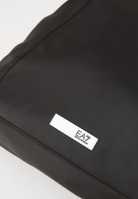 Borsa di stoffa nera con una superficie liscia, design pulito e un'etichetta bianca con il marchio "EA" nell'angolo.