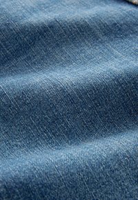 Denim tkanina u srednje plavoj nijansi, koja prikazuje teksturiranu površinu s suptilnim varijacijama i klasičnim twill uzorkom.
