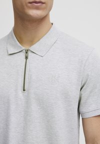 Grå polo shirt med blød tekstur, der har en lynlås-i-kraven og broderet logo på venstre bryst. Korte ærmer og tætsiddende pasform.