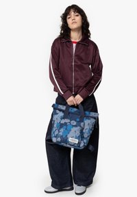 Bolso tote con estampado floral en tonos azul y gris, con asas superiores y un bolsillo frontal. El modelo lleva una chaqueta de cremallera color burdeos y unos jeans oscuros holgados.
