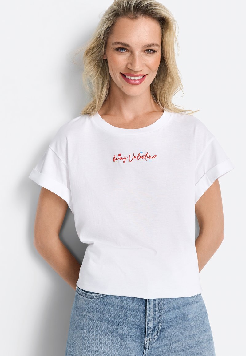 T-shirt blanc à manches courtes avec des poignets retroussés et un col rond, présentant un design de texte "sois mon Valentine" en rouge et bleu sur le devant.