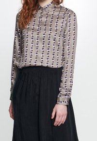 Långärmad blus i en tan-färg med en knappad krage och vertikala rader av lila cirklar. Slät tyg, matchad med en svart kjol.