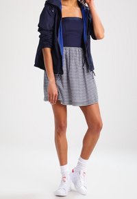 Kvinna i marinblå jacka, strapless marinblå topp, vit och marinblå randig kjol, vita strumpor och vita sneakers som står mot en vit bakgrund.