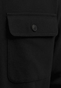 Camicia nera in tessuto con tasca frontale con chiusura a bottone. Texture liscia, vestibilità su misura e elementi di design minimalisti.